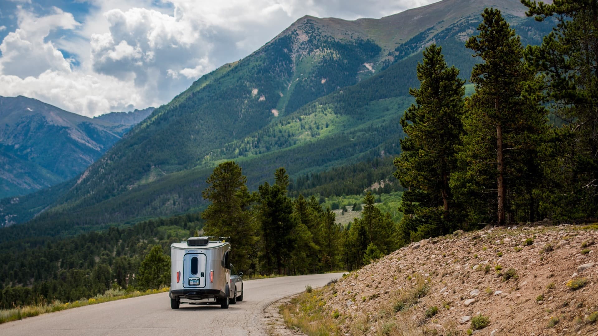 Small travel trailer driving on mountain road with trees and clouds