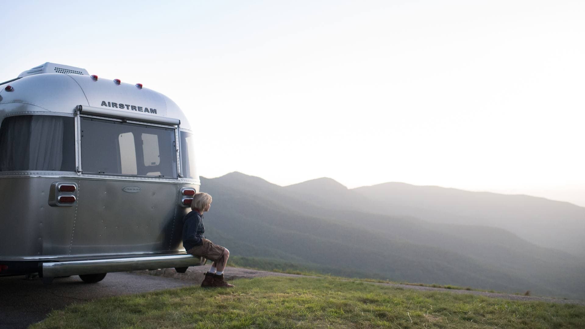Woman sitting alone on trailer step overlooking mountainous landscape at sunset