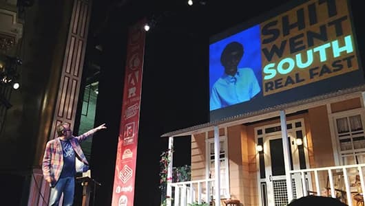 Speaker on stage under large outdoor video screen at nighttime event