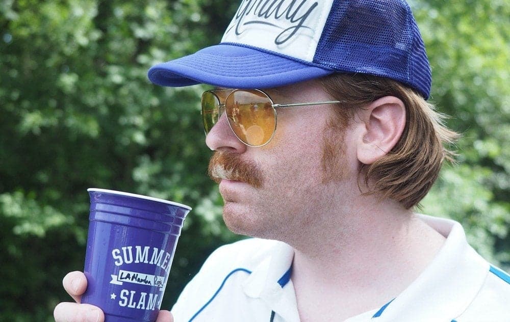Man wearing sunglasses and trucker hat holding beverage at outdoor event