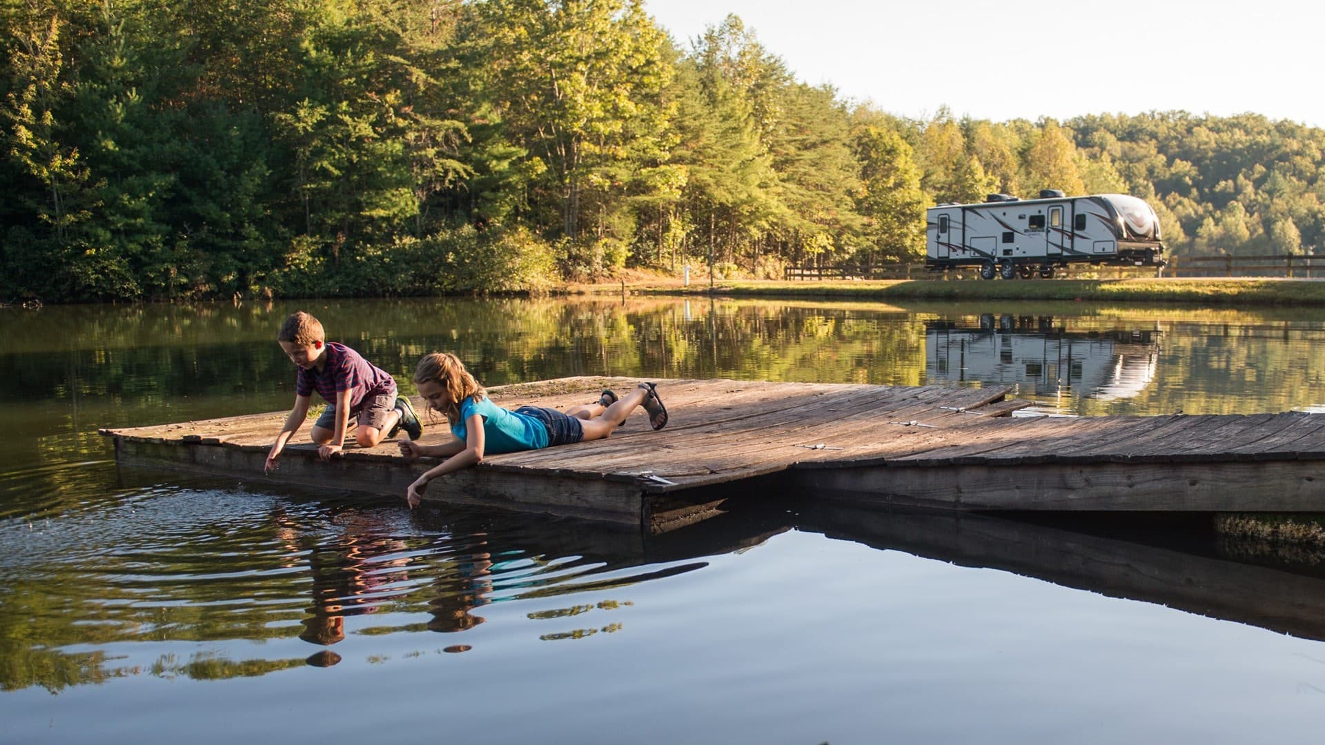Woman and child lounging at water's edge with travel trailer and forest visible